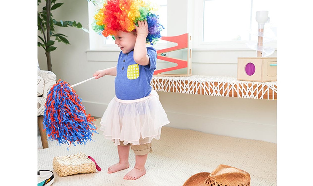 Toddler playing dress up with a clown wig, skirt, and pom pom surrounded by items by Lovevery