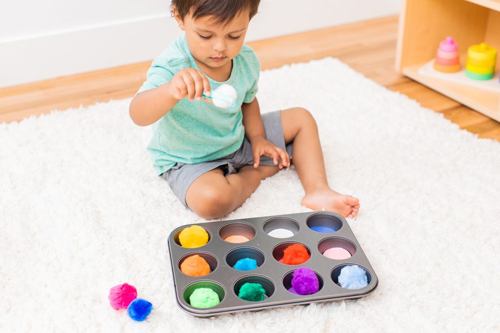 Young child picking up colorful pom poms from a muffin tin