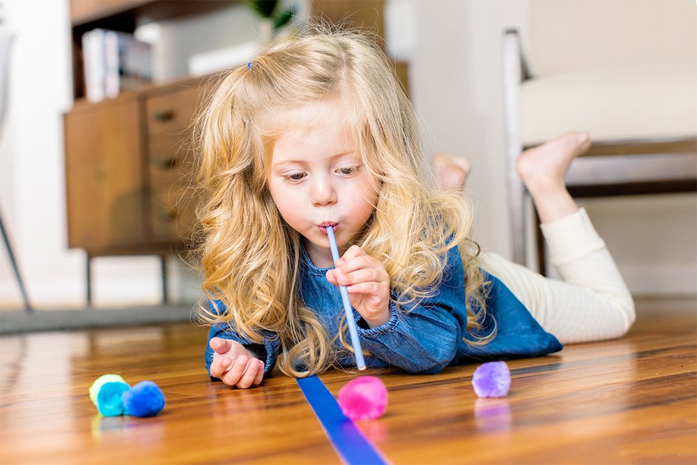 Young child blowing through a straw moving a pom pom
