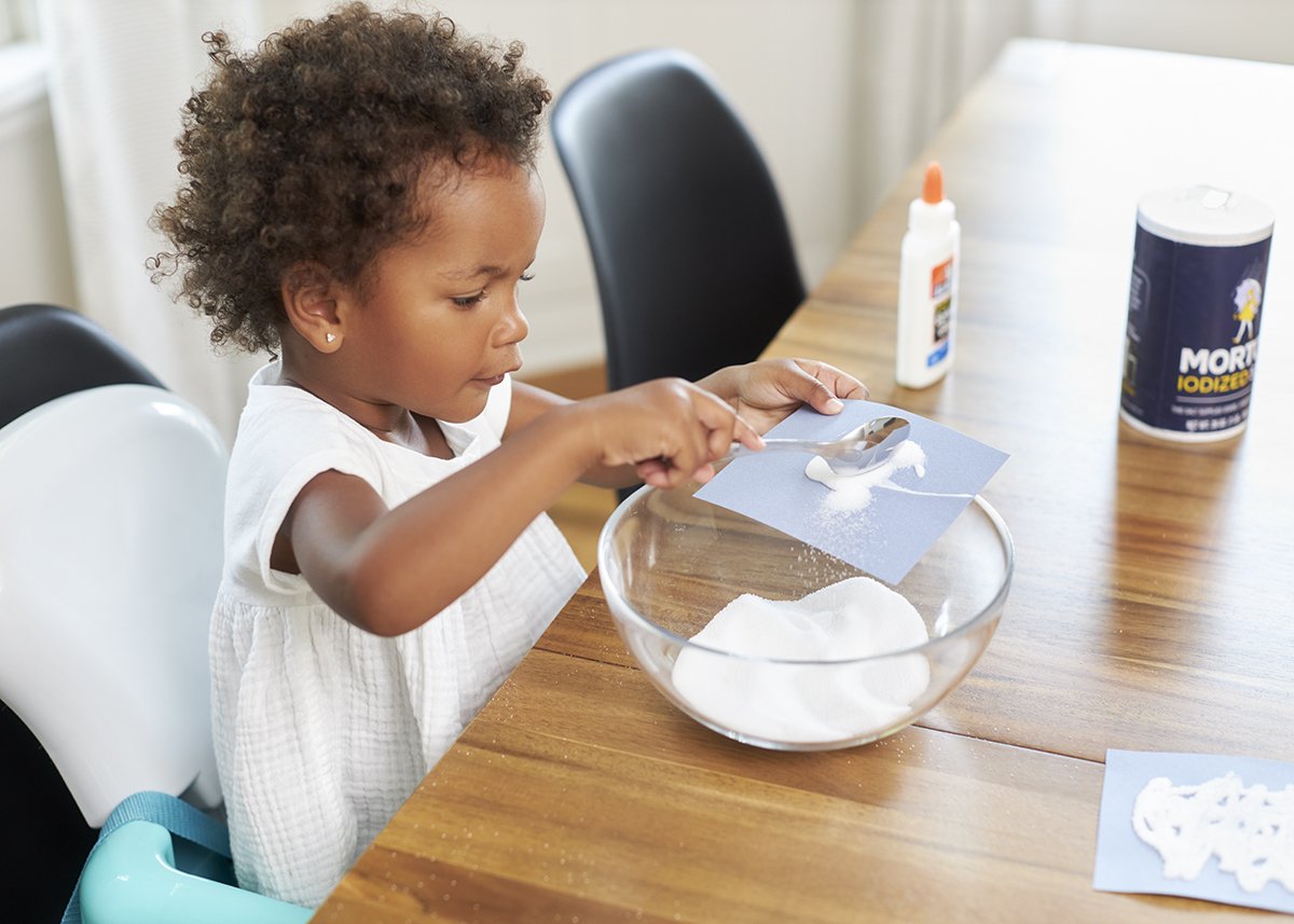 Young child holding a piece of paper with glue on it and pouring salt on top.